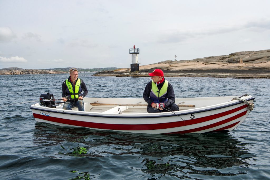 Två personer i en fiskebåt med en fyr i bakgrunden.